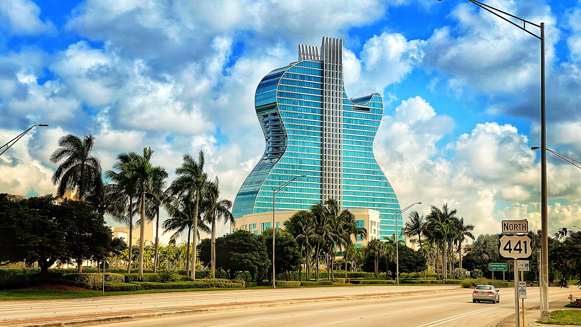 The guitar-shaped glass tower of the Seminole Hard Rock Hotel rising above palm trees beside US 441 under a cloudy blue sky