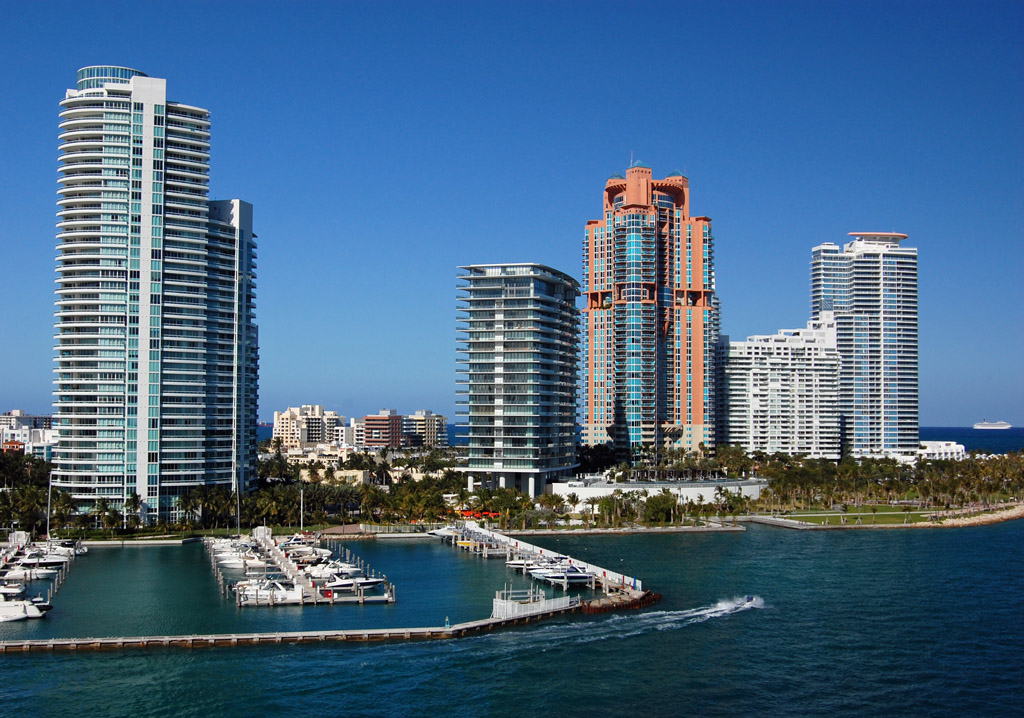 High-rise condominium towers at the southern tip of Miami Beach rising above palm trees, with a marina of white boats in the foreground and a cruise ship far out to sea
