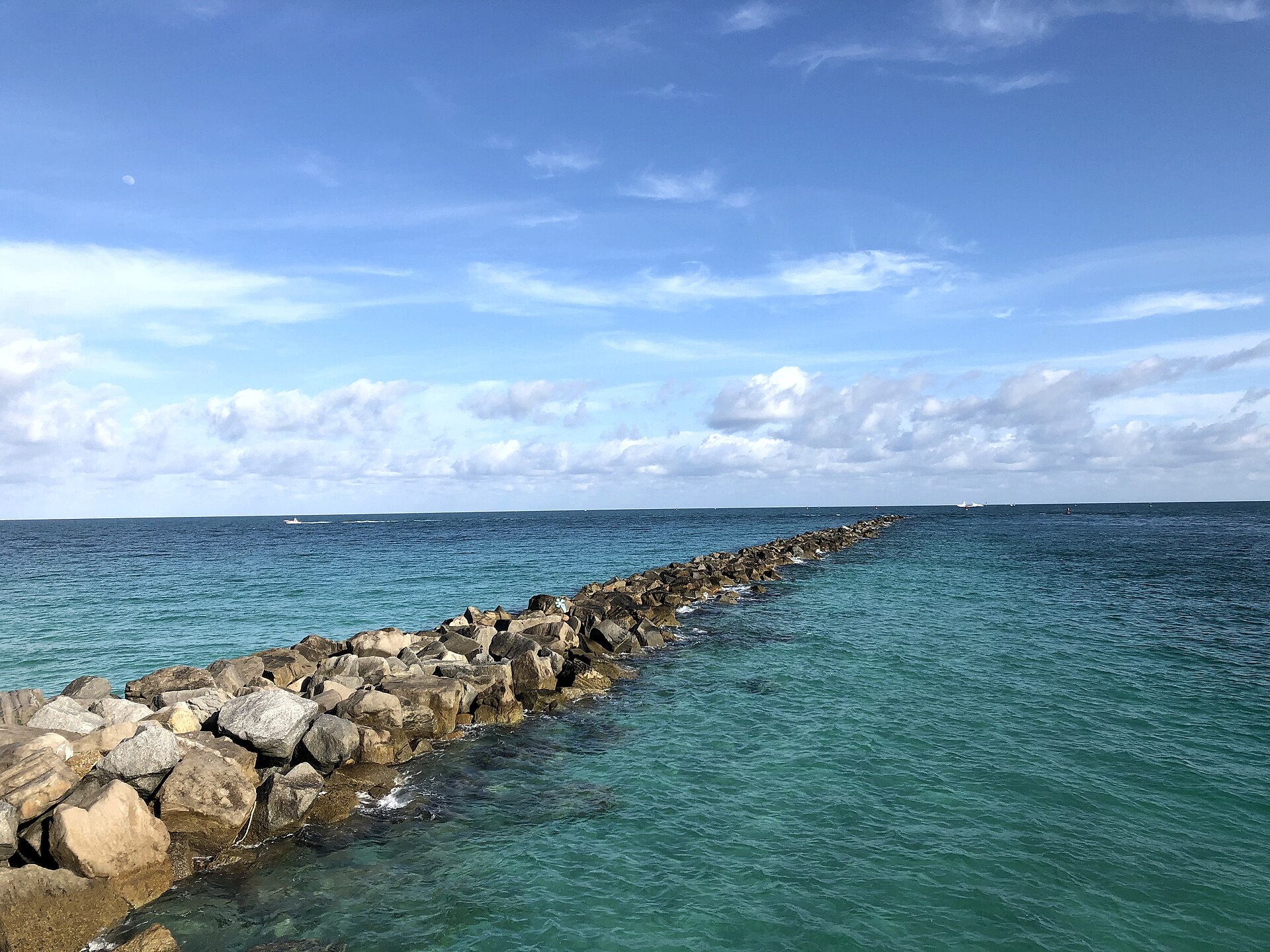 A long rock jetty running straight out from the shore into clear turquoise water under a blue sky with a faint daytime moon
