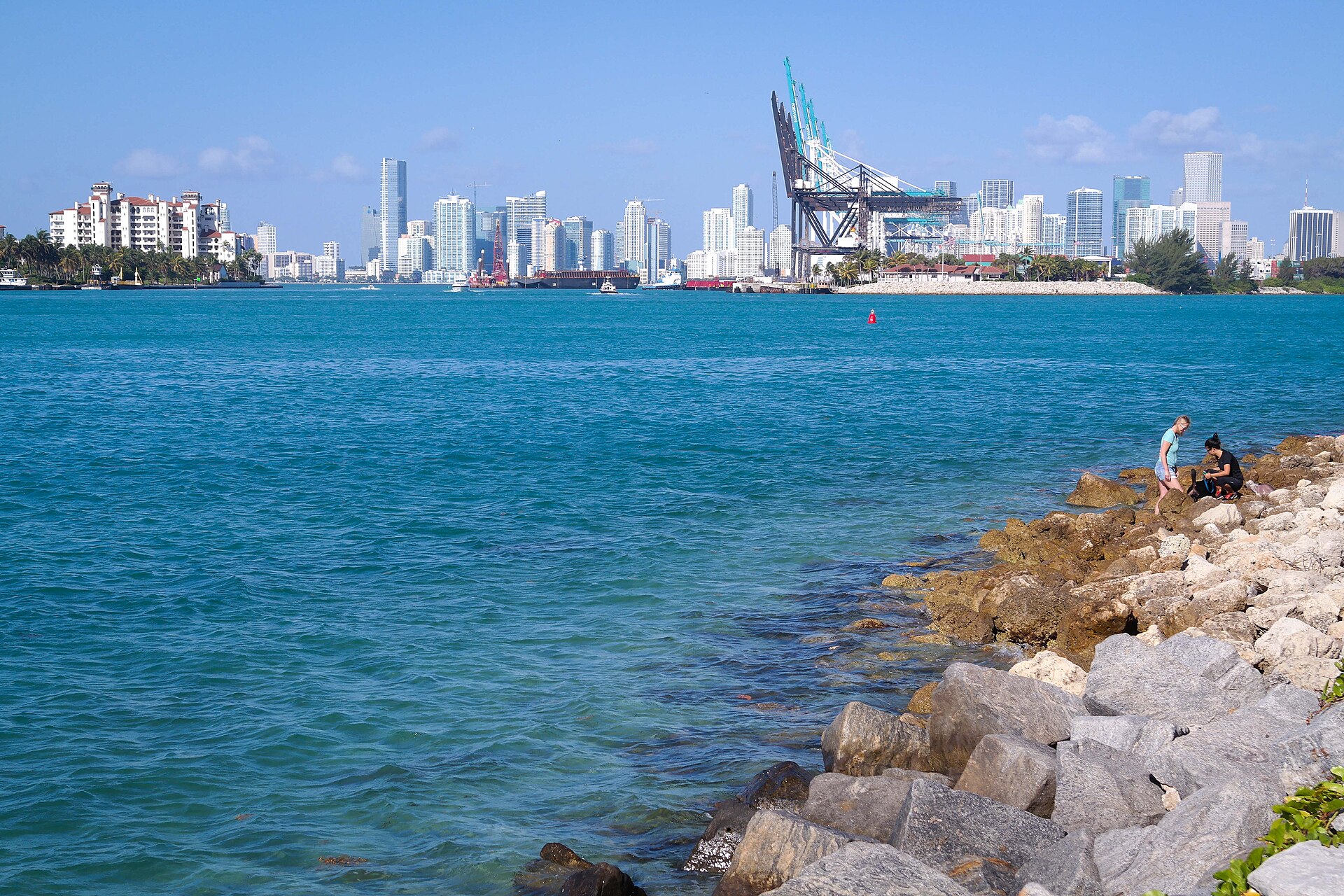 View from the rocks at South Pointe Park across turquoise water to Fisher Island's red-roofed buildings, the PortMiami cranes and the downtown Miami skyline