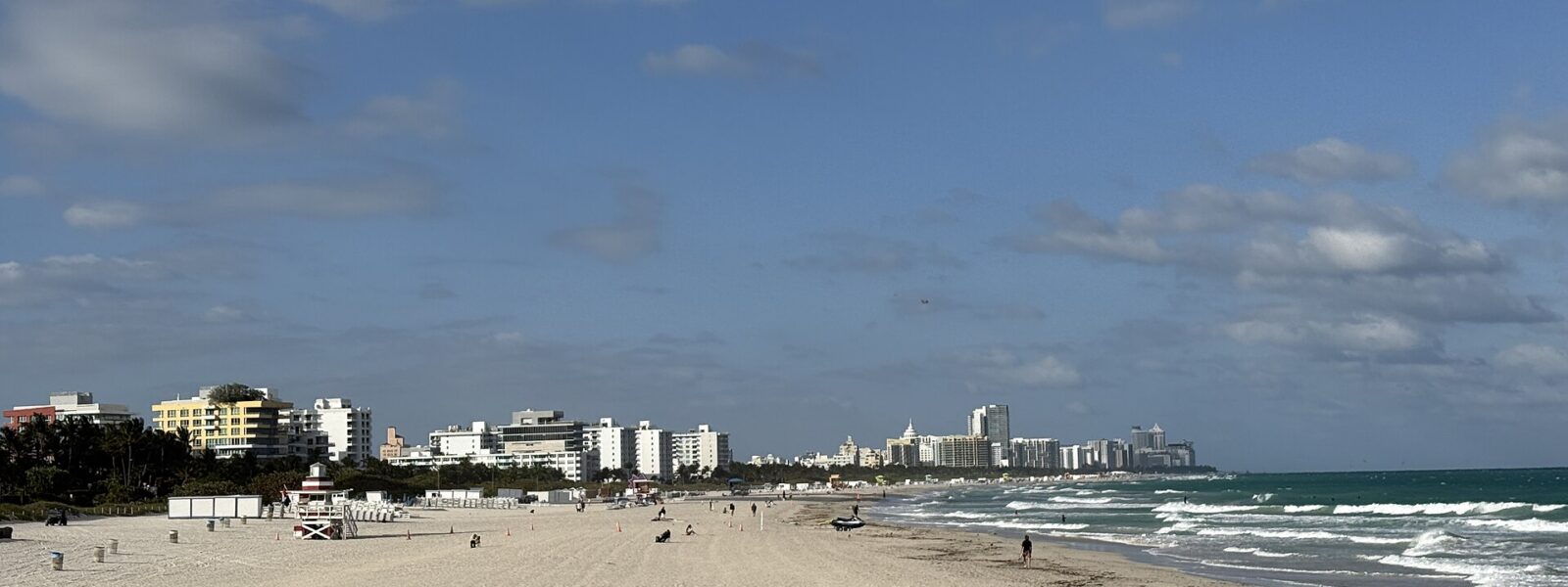 The wide sand of South Pointe Beach seen from the pier, with a red and white lifeguard tower and the South Beach hotels stretching north under a blue sky