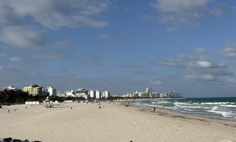 The wide sand of South Pointe Beach seen from the pier, with a red and white lifeguard tower and the South Beach hotels stretching north under a blue sky
