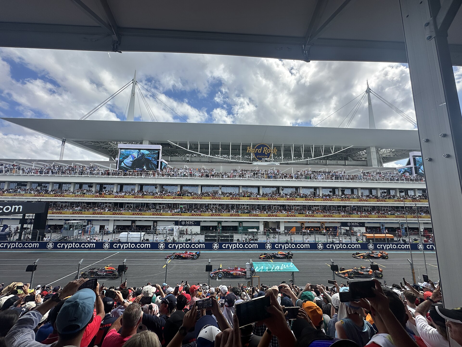 Spectators in a covered grandstand watch the Miami Grand Prix starting grid, with Hard Rock Stadium rising behind the hospitality balconies.