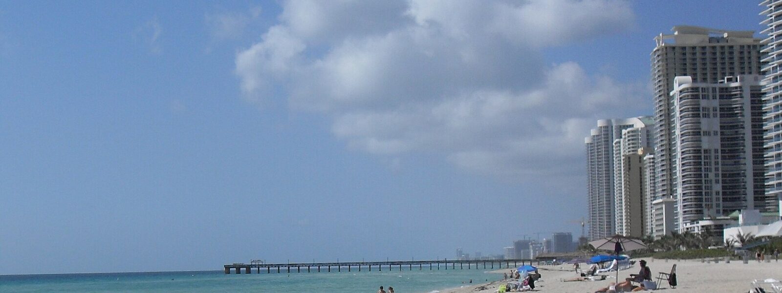 Wide pale sand at Sunny Isles Beach with a few swimmers in turquoise water, a long fishing pier reaching out to sea and high-rise condominium towers along the shore