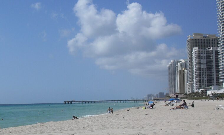 Wide pale sand at Sunny Isles Beach with a few swimmers in turquoise water, a long fishing pier reaching out to sea and high-rise condominium towers along the shore
